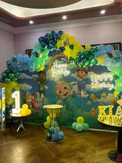 A Cocomelon first birthday party with a large character backdrop. The decoration features a balloon arch, a "Happy Birthday" neon sign, and marquee letters for the name.