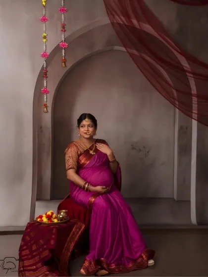 A beautiful solo portrait of the mama-to-be, seated gracefully. The magenta saree and traditional props create a rich, cultural feel.