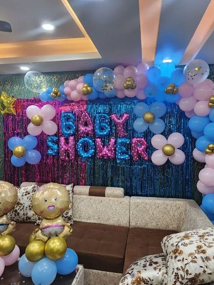 A cozy living room setup for a baby shower. The pink and blue balloon arch over a shimmering backdrop, along with flower-shaped balloon clusters, adds a festive touch to the home.