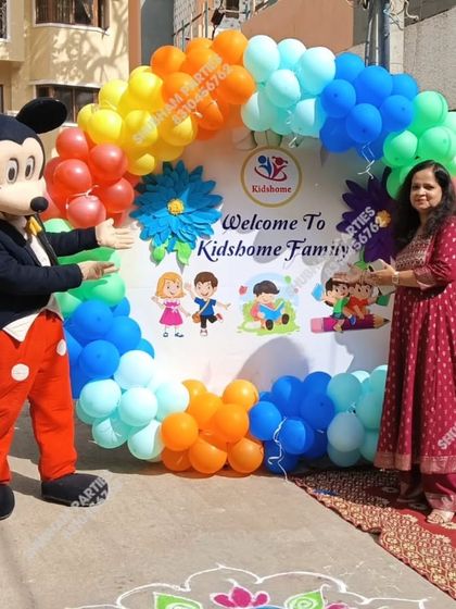 A Mickey Mouse mascot greeting guests at a school event with a rainbow balloon ring backdrop.