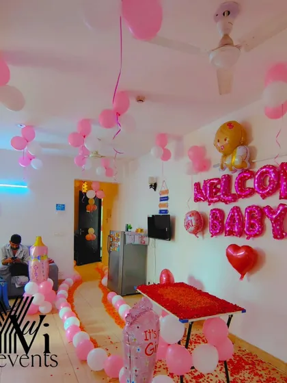 A full room decoration for a "Welcome Baby" girl party. The setup includes a balloon pathway, a decorated table, and foil balloons on the wall.