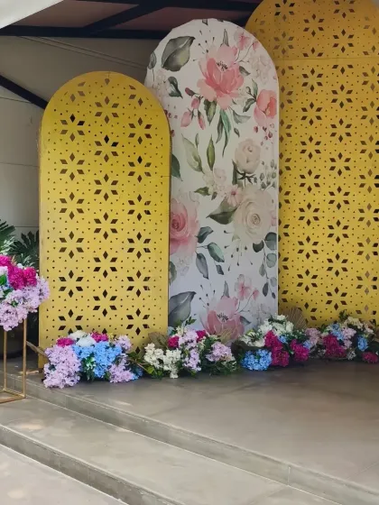 A modern and cheerful photo backdrop for an engagement party. The design uses three arched panels, two with yellow jaali-cut patterns and a central one with a watercolor floral print, accented with colorful flowers.