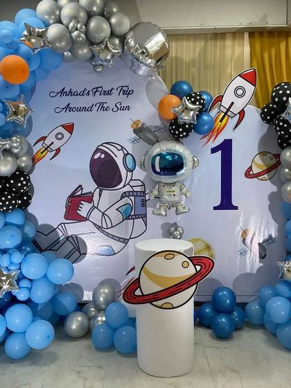 "First Trip Around The Sun" space-themed party. This decoration features a custom astronaut backdrop, a balloon arch with planet and rocket foils, and a light-up number '1'.
