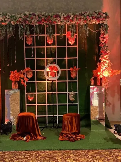 A creative photo booth or seating area for a Mehndi function. The setup includes a white grid backdrop, hanging floral elements, and velvet-draped stools on a patch of green.