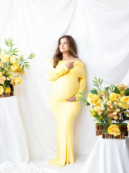 A sunny and cheerful studio set. The expectant mother in her yellow gown is framed by beautiful arrangements of yellow flowers, creating a bright and happy scene.