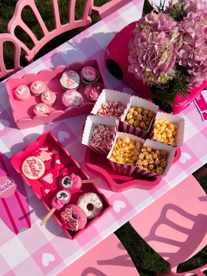 An overhead view of the Barbie-themed birthday party setup. The table is a sea of pink, featuring custom trays with themed desserts, gourmet popcorn, and even a Barbie surfboard prop.