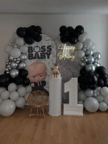 A stylish Boss Baby first birthday in black, white, and silver. The setup includes a custom backdrop, a "1" marquee number, and modern balloon arches.