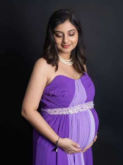 A serene close-up of the mom-to-be cradling her bump. The soft lighting beautifully illuminates her features as she looks down with a gentle expression, wearing an elegant purple gown.