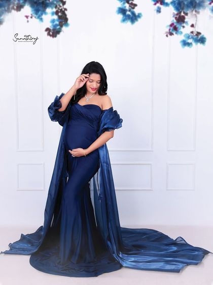 A stylish indoor portrait of a mother-to-be in a deep blue gown. The simple white paneled background and floral accents create a clean and elegant look.