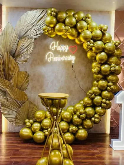 I design elegant and luxurious setups for milestone anniversaries. This first-anniversary decoration features a stunning all-gold chrome balloon ring, a custom "Happy Anniversary" neon sign, large marquee numbers, and decorative dried palm leaves for a modern, sophisticated look.