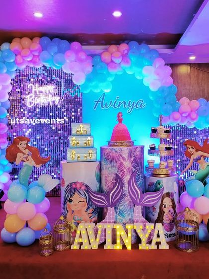 A magical Mermaid-themed birthday party. The setup features a blue sequin backdrop, character cutouts of Ariel, and balloon arrangements in ocean colors to create an underwater feel.