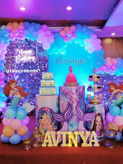 A magical Mermaid theme for Avinya's birthday. The decor features a blue shimmer wall, mermaid tail props, character cutouts, and a pastel balloon arch.