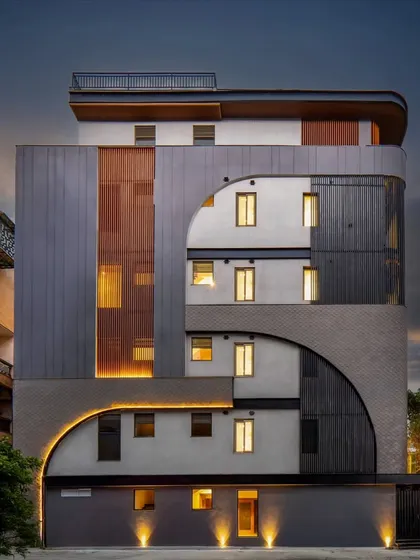 The Patel Nagar builder floor at night, where accent lighting highlights the symmetrical curves and material palette. The design combines aluminum louvers, concrete, and textured mortar in grey-black hues for a grounded presence.