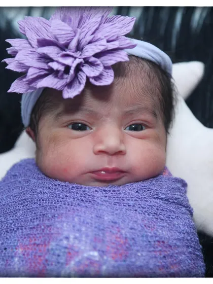 A beautiful close-up of a newborn baby girl with a lovely purple flower headband, wrapped in a matching purple swaddle.