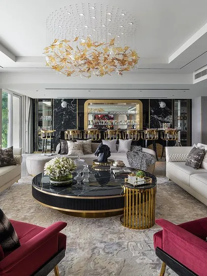 A vibrant and luxurious living room featuring bold red armchairs, a black marble coffee table, and a mix of patterned and textured cushions. The space is designed for entertaining, with a fully stocked bar visible in the background.