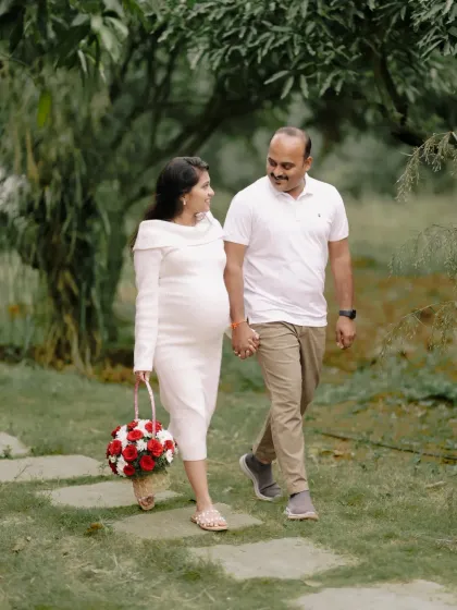 An expecting couple enjoys a quiet walk along a garden path. The mother-to-be holds a basket of flowers, adding a lovely detail to this candid moment of connection and conversation.