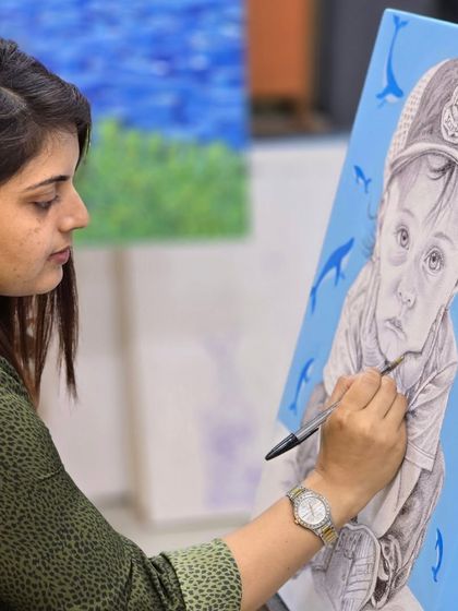 A student carefully works on her portrait drawing, using a ballpoint pen to add details to the child's clothing and expression.