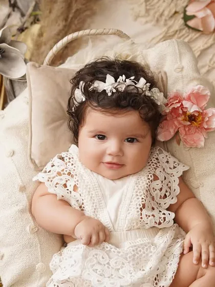 A beautiful baby girl in a white lace romper and floral headband smiles sweetly while lying in a basket, surrounded by soft fabrics and flowers.