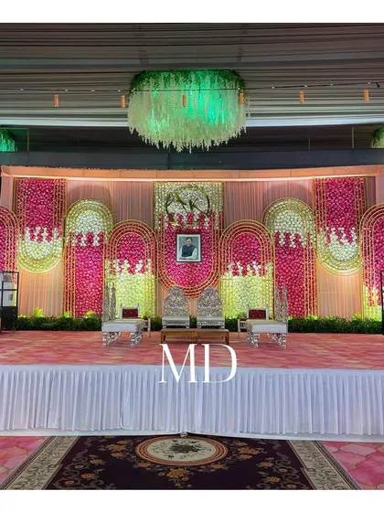 A different perspective of a floral stage setup, suitable for a Vidhi or engagement ceremony. The backdrop is made of arched panels filled with pink and yellow flowers, creating a vibrant and celebratory look.