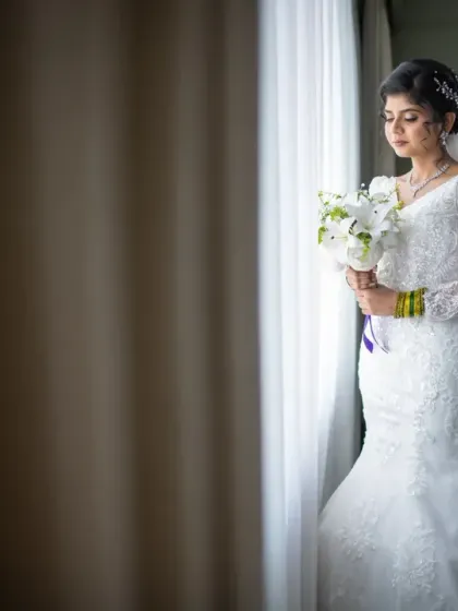 A full-length bridal portrait showcasing the bride's stunning mermaid-style gown and long veil. The soft window light adds a touch of romance.