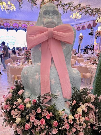 A whimsical, life-sized gorilla statue wearing a giant pink bow, surrounded by a bed of pink roses. This is an example of a bold and unexpected bespoke prop.