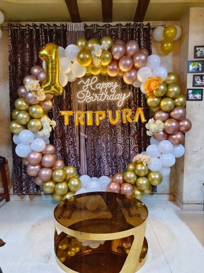 A premium first birthday setup for Tripura, featuring a full balloon ring in white, gold, and rose gold. The design includes a neon sign and a foil number '1' balloon.