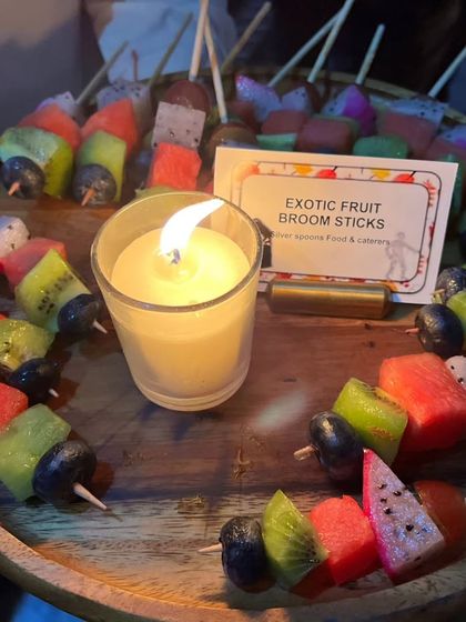 'Exotic Fruit Broom Sticks' offer a healthy and refreshing option for a Halloween party. The colorful fruit skewers are a vibrant contrast to the spooky decor, all lit by a candle.