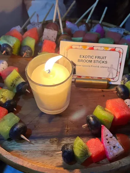 'Exotic Fruit Broom Sticks' offer a healthy and refreshing option for a Halloween party. The colorful fruit skewers are a vibrant contrast to the spooky decor, all lit by a candle.