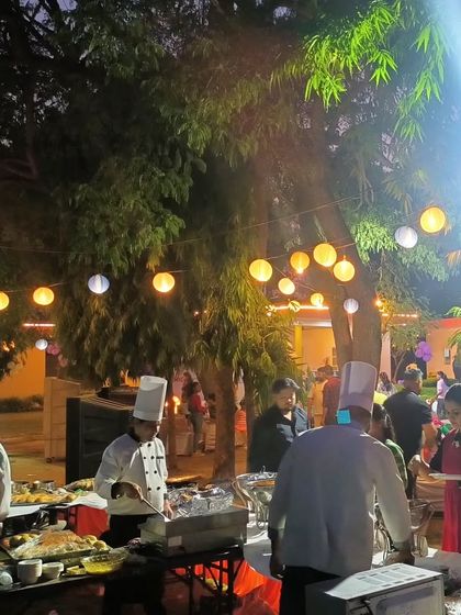 An atmospheric shot of our live cooking stations at an evening garden party. The warm lighting from the lanterns and the active chefs create a vibrant and inviting scene for guests.