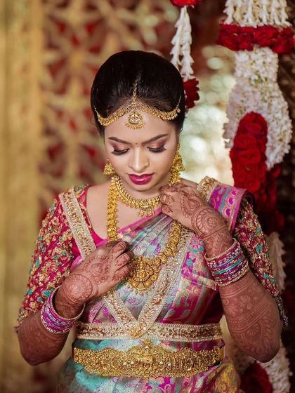 The bride adjusting her necklace, a moment that shows the complete look with her traditional gold jewelry. The makeup is designed to enhance, not overpower, these beautiful elements.
