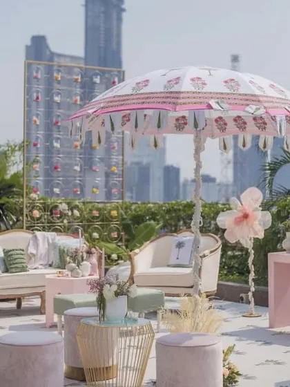 A chic and modern rooftop Mehendi in an urban setting. The pastel pink and white decor, elegant seating, and custom-printed umbrella create a sophisticated and stylish ambiance with a stunning city skyline view.
