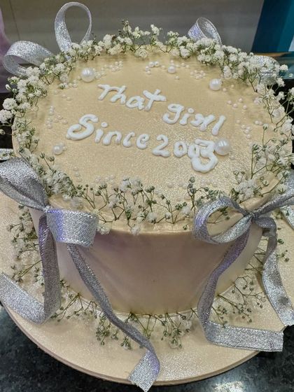 An elegant and dreamy cake with the message "That girl since 2003." It's decorated with delicate baby's breath and silver ribbons, creating a soft and ethereal look.