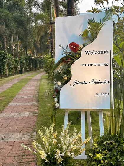 A modern and elegant welcome sign for a lakeside wedding. The organic shape of the sign and the tropical floral arrangement blend seamlessly with the natural surroundings.