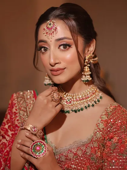 A close-up of this model's timeless elegance. The makeup is classic bridal, with defined eyes, glowing skin, and a neutral lip that complements the traditional red and green jewelry.
