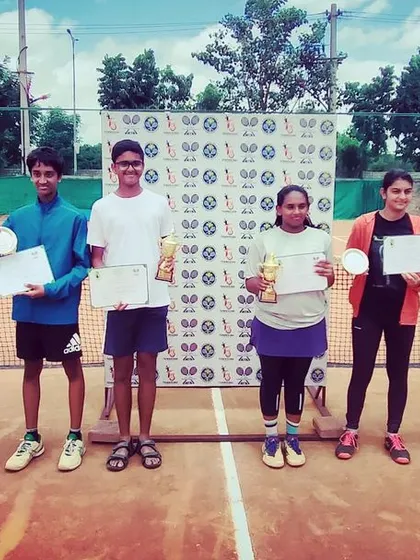 A group photo of all the winners and runners-up from the AITA TS7 Under-14 tournament, celebrating their achievements with trophies, medals, and certificates.