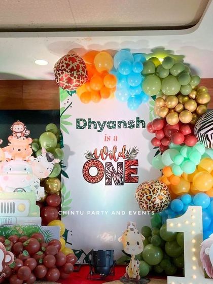 A 'Wild One' first birthday party with a jungle safari theme. The setup includes a custom backdrop, animal print balloons, character cutouts, and a light-up marquee number '1'.
