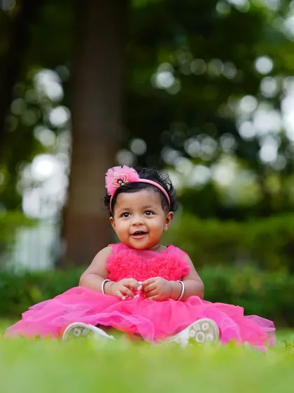 A beautiful portrait of a baby girl in a bright pink dress, sitting on the green grass. The natural background makes the colors pop in this outdoor shot.