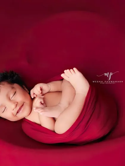 A sweet, sleepy smile captured from the side. The red wrap and heart prop create a classic and emotive newborn photograph.