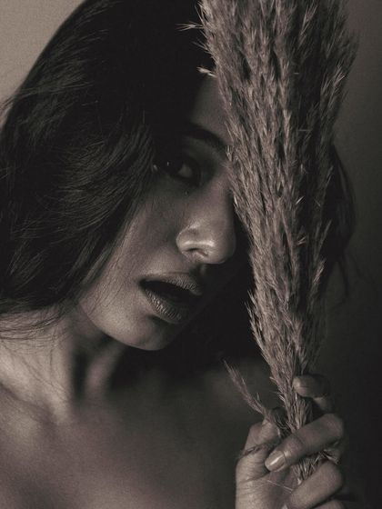 The texture of the pampas grass contrasts with the softness of the model's skin in this moody black and white close-up. The lighting is dark and dramatic, creating a sense of mystery.