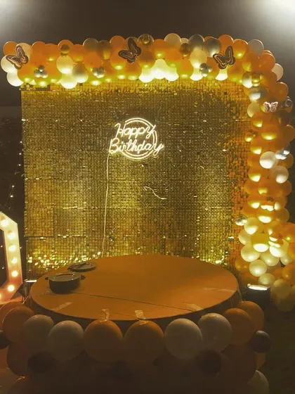 A golden-themed 20th birthday party setup. The decor includes a gold sequin backdrop, a balloon arch, and large marquee numbers, creating a glamorous photo corner.