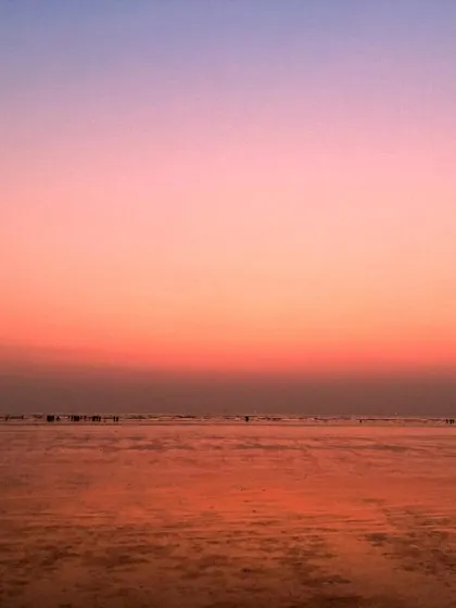The crescent moon is visible in a gradient sky of orange and pink, above a calm beach in Mumbai at low tide.