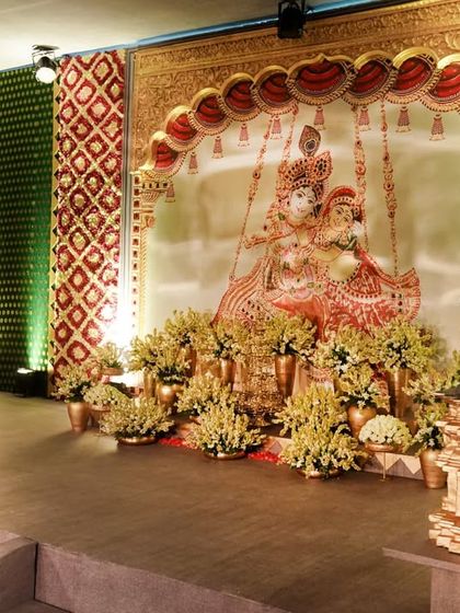 A side stage within our temple-themed wedding, featuring a large Tanjore-style painting of Radha and Krishna. The area is flanked by intricately carved pillars and adorned with yellow flowers, serving as a beautiful photo backdrop and lounge space.