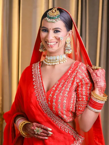 A bright and cheerful bridal portrait featuring a smiling model in a red embroidered outfit. This image showcases a softer bridal makeup style with a natural glow, complemented by a choker-style necklace and traditional red and gold bangles.