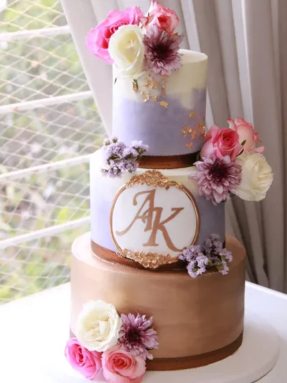 An elegant three-tiered anniversary cake featuring a blend of lavender ombre, a rose gold bottom tier, and a custom monogram. The cake is finished with my smooth colored ganache and fresh flowers.