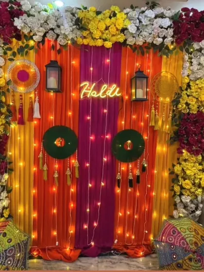 This Haldi backdrop combines colorful drapes with a custom "Haldi" neon sign. The decor is enhanced with dreamcatchers, lanterns, and traditional printed cushions for a bohemian-chic vibe.