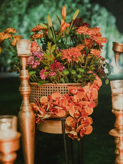 A beautiful arrangement of orange orchids and chrysanthemums in a cane planter, surrounded by the warm glow of candles in tall, elegant holders. This adds a touch of warmth and nature to the glam setup.