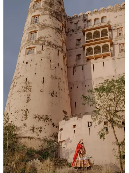 A wide-angle shot that captures the scale of the venue and the solitude of the bride. Amruta, in her beautiful red lehenga, stands against the towering walls of a historic fort, a queen in her castle.