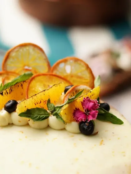 A close-up of a beautifully decorated orange cake, garnished with dehydrated orange slices, fresh basil, and blueberries. A perfect cake for a sunny celebration.
