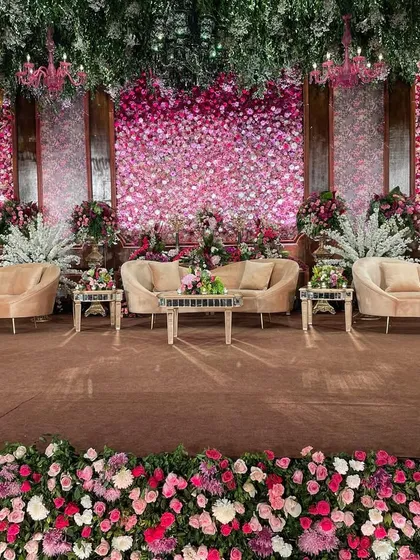 A regal reception stage featuring walls of pink and red flowers against rich wood paneling. The plush velvet sofas and intricate floral border complete this luxurious and opulent look.