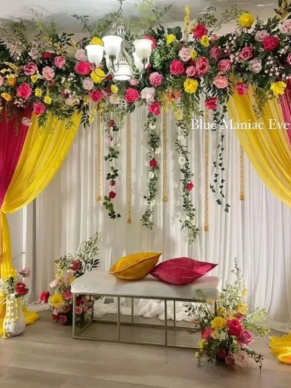 An elegant Mehendi setup with a ceiling floral installation. The pink and yellow drapes frame a simple bench, creating a beautiful and sophisticated look.